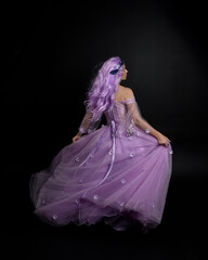 Full length portrait of girl wearing long purple fantasy ball gown with crown and pink hair, standing pose with back to the camera  against a studio background.