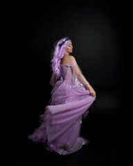 Full length portrait of girl wearing long purple fantasy ball gown with crown and pink hair, standing pose with back to the camera  against a studio background.