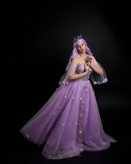 Full length portrait of girl wearing long purple fantasy ball gown with crown and pink hair, standing pose holding a wand  against a studio background.