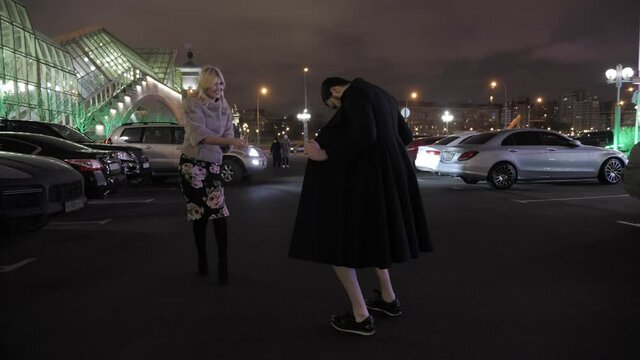 beautiful lady laughs looking at funny exhibitionist in black coat and sunglasses on parking lot at dark night