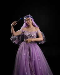 Close up portrait of girl wearing long purple fantasy ball gown with butterfly crown and pink hair, against a  dark studio background.
