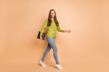 Fototapeta na wymiar Photo of girl walk hold computer device wear green pullover jeans sneakers isolated beige color background