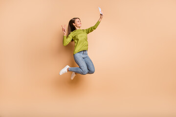Fototapeta na wymiar Profile photo of girl jump hold phone make selfie show v-sign wear green pullover jeans sneakers isolated beige color background