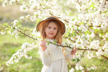 A sweet happy little girl in a white dress and a straw hat in spring near a flowering tree