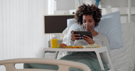 Afro-american woman patient sitting in bed with tray of food and using digital tablet at lunch