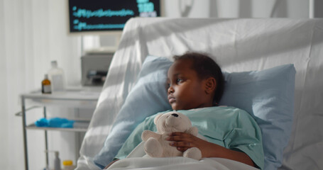 Portrait of sick african girl hugging teddy bear lying in hospital bed