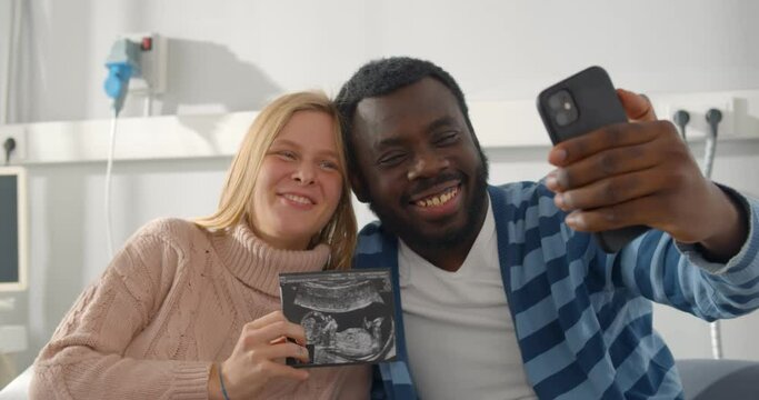 Happy multiracial married couple holding ultrasound picture of baby and taking selfie