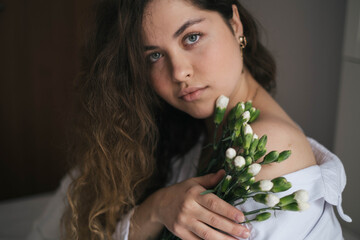 close up of a pretty caucasian woman with curly hair smiling with a bouquet flower sit on bed looking at camera. Concept of spring, feminity, beauty, spa. Womans day, birthday.