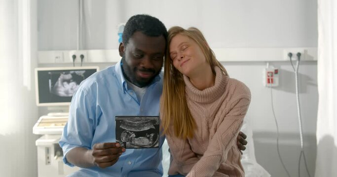 Happy multiethnic couple holding ultrasound scan of their baby sitting in clinic office
