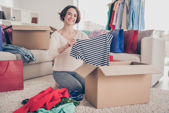 Full size photo of happy positive young woman hold clothes unpack box move apartment sit floor inside home