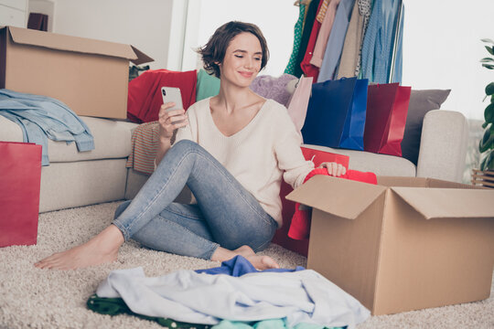 Full length photo of happy beautiful young woman look box second hand clothes hold phone sit floor inside house