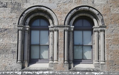 Two decorative, old and symmetrical arched stone windows in a grey wall. 