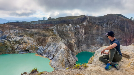 Man sitting at the volcano rim and watching the Kelimutu volcanic crater lakes in Moni, Flores, Indonesia. Man is relaxed and calm, enjoying the view on turquoise lakes. Lakes have different colours