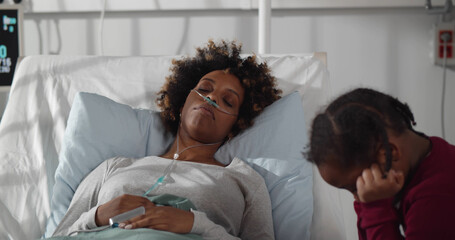 Little upset african girl sitting at bedside of sick mother in hospital ward