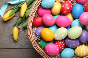 Fototapeta na wymiar Colorful Easter eggs in wicker basket and tulips on wooden table, flat lay