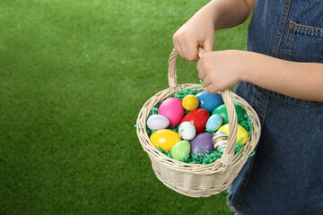 Little girl with basket full of Easter eggs on green grass, closeup. Space for text