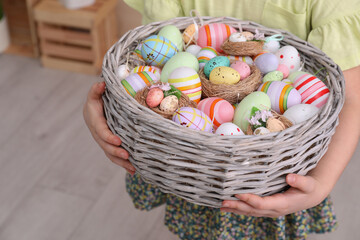 Little girl holding wicker basket full of Easter eggs indoors, closeup