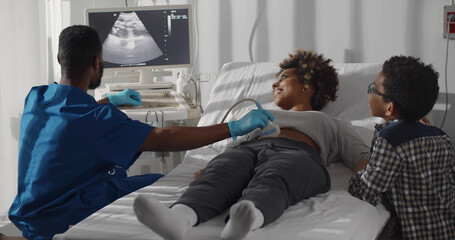 Little african kid standing near mother having ultrasound at doctor