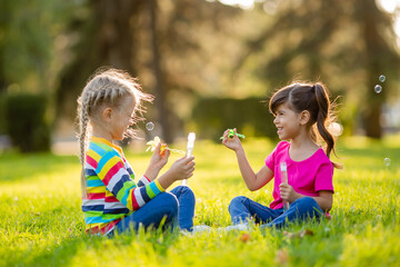 Two little blonde girls and brunette summer sit on the lawn blowing soap bubbles. European and...