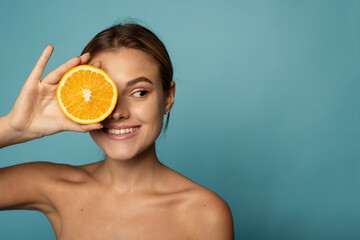 Attractive caucasian female with clean skin laughing with closed eyes and showing half of fresh orange while advertising benefits of vitamin C