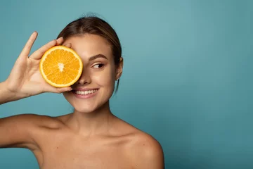 Fotobehang Attractive caucasian female with clean skin laughing with closed eyes and showing half of fresh orange while advertising benefits of vitamin C © etonastenka