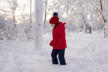 Little Caucasian girl in a red knitted hat and jacket walks in the snow in the park in winter. Winter fun for children and frosty air.