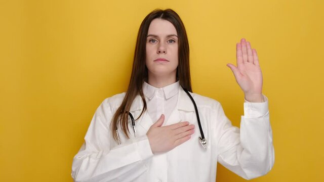 Honest young female doctor, intern giving pledge, raising one arm and hold hand on heart while promise, oath to patient, isolated on yellow background. Healthcare workers and preventing virus concept
