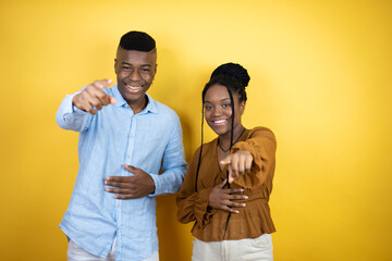 Young african american couple standing over yellow background laughing at you, pointing finger to the camera with hand over body, shame expression
