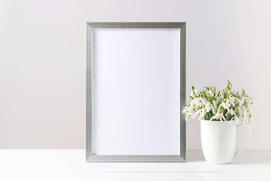 Greeting Hello spring card mockup or silver photo frame in modern scandinavian style with bouquet snowdrop flowers in the ceramic cup on wooden table. White background with copy space
