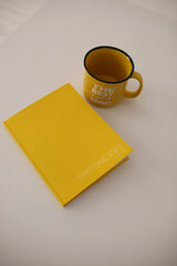 A yellow mug with the words the best is yet to come and a yellow notebook on a white table top view. A mug and a notebook on a white table in the color of the year.