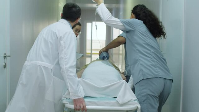 Rear view of team of paramedics using bag-valve-mask and IV bag while pushing emergency patient on gurney through hospital corridor