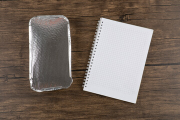 Closed aluminum foil plate of sushi rolls and notebook on wooden surface