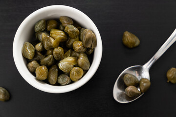 Top view of a bowl of green capers and a spoon isolated on a black background. 