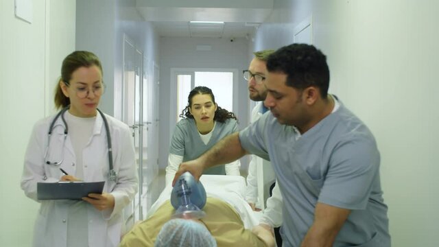Tilt up shot of team of multiethnic paramedics giving bag-valve-mask ventilation to emergency patient and pushing gurney towards the camera through hospital hallway