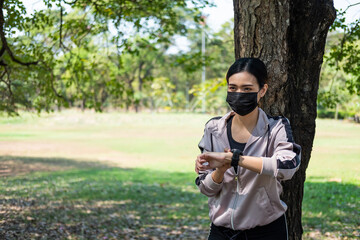 Selective focus at face of young beautiful Asian women wearing surgical face mask using smart watch to track activity before exercise or running at the park in the morning. New normal lifestyle.