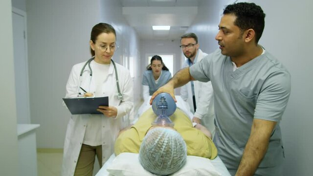 Group of multiethnic doctors giving bag-valve-mask ventilation to emergency patient and pushing gurney along hospital hallway