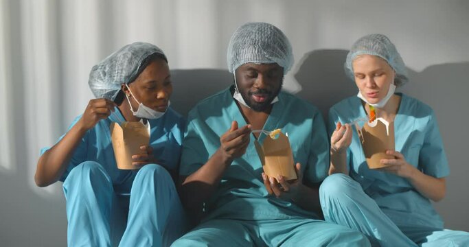 Diverse healthcare workers in scrubs uniform talking and eating takeaway food sitting on floor