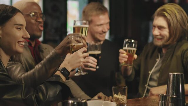 Slow-motion medium shot of cheerful young company of multi-ethnic friends having fun together at local pub, toasting beer glasses and laughing