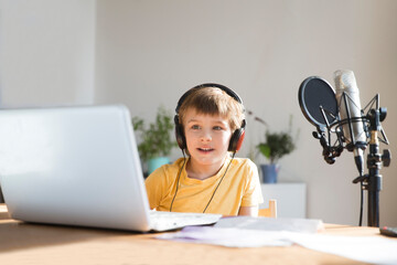 Smiling schoolboy using headphones in front of microphone talking and recording podcast in home...
