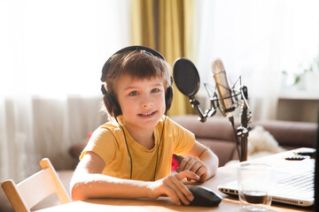 Smiling schoolboy using headphones in front of microphone talking and recording podcast in home...