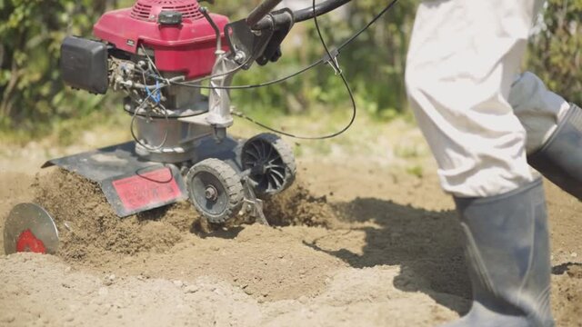 Tiller - motor cultivator working on garden, cultivating soil for new season. Farmer in light roba and rubber boots cultivates the soil. Footage Full HD. Very close view