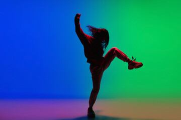 Naklejka na ściany i meble Modern. Stylish sportive girl dancing hip-hop in stylish clothes on colorful background at dance hall in neon light. Youth culture, movement, style and fashion, action. Fashionable bright portrait.