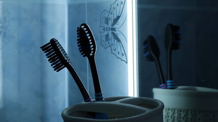 two black toothbrushes in a cup on a shelf in the bathroom near the mirror in dark blue dim...