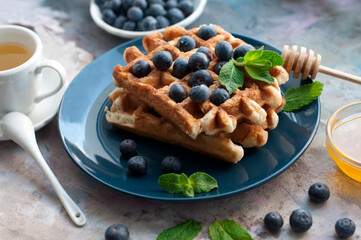 Belgian waffles with blueberries, honey and mint on a blue plate. Breakfast with a cup of tea and sweet waffles