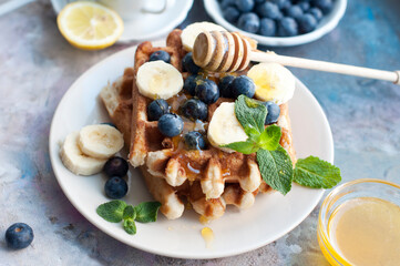 Belgian waffles with blueberries, bananas, honey and mint on a white plate. Breakfast with a cup of tea with a lemon and sweet waffles. 