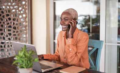 Beautiful mature african woman working with laptop while talking on a call with smartphone