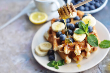 Belgian waffles with blueberries, bananas, honey and mint on a white plate. Breakfast with a cup of tea with a lemon and sweet waffles. Honey flowing from a wooden honey stick
