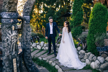 Young beautiful couple walks in a green park in summer near the stones. The bride is in a white dress with a long train, the groom is in a suit.