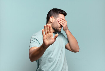 young handsome adult man covering face with hand and putting other hand up front to stop camera,...