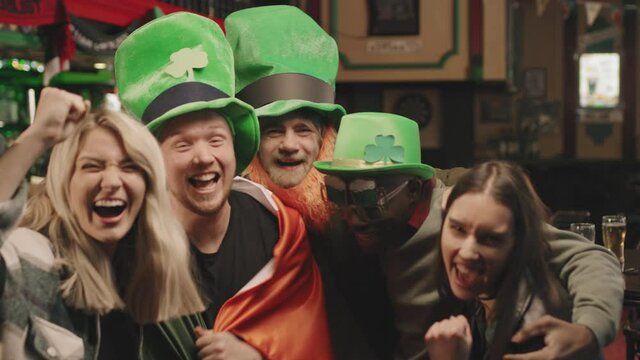 Slow-motion medium shot of group of young multi-ethnic friends wearing handmade green hats with clover poster cheering to camera celebrating St Patricks Day together in local Irish pub